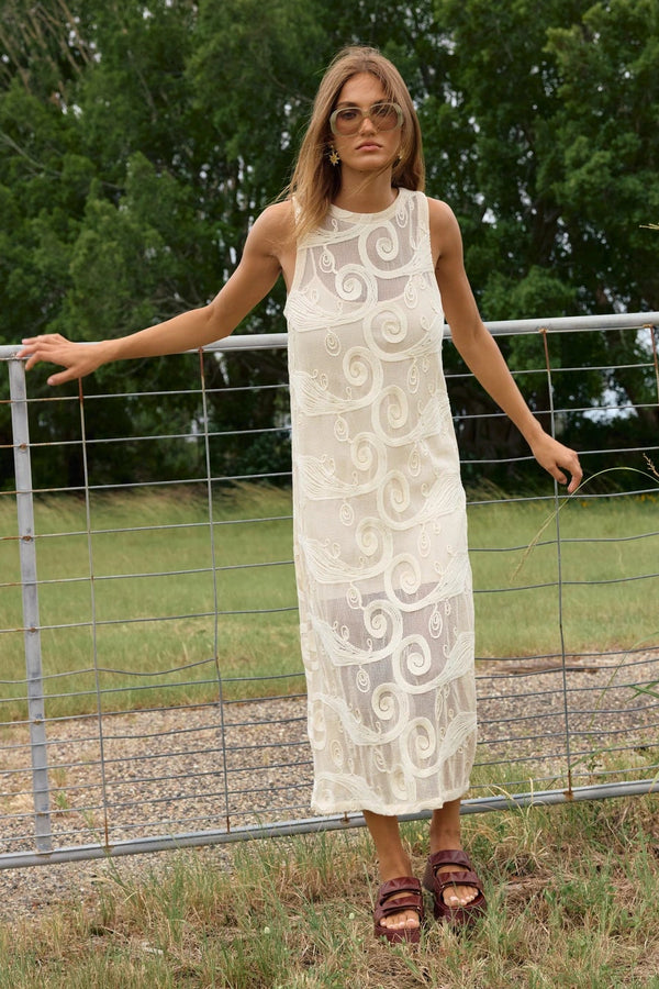 A woman wearing a sleeveless cream lace midi dress with circular patterns and brown sandals, standing outdoors in front of a wire fence and green trees.