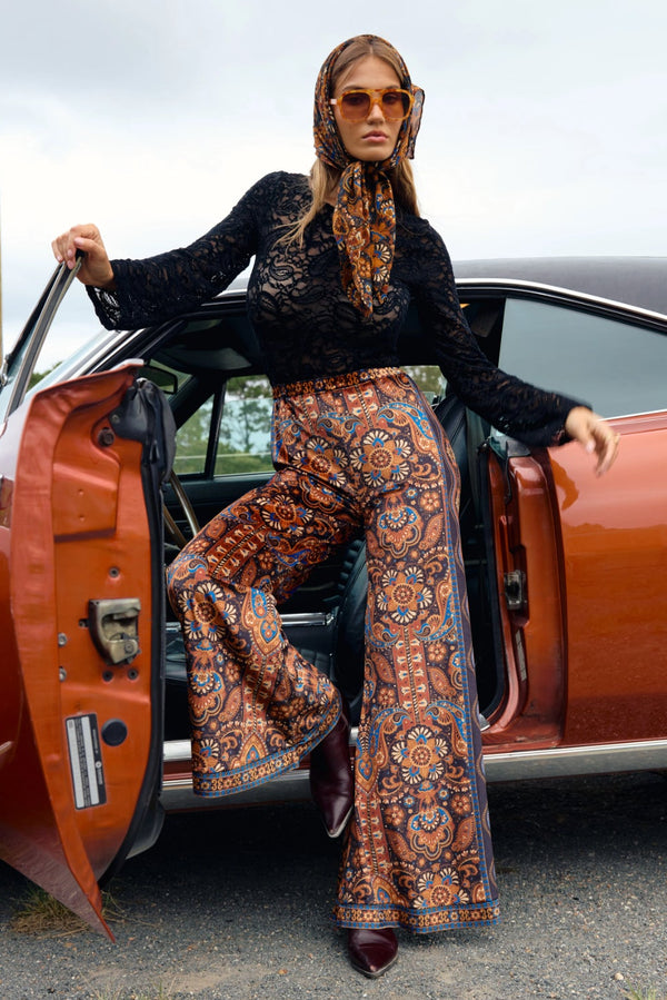 Person in a black top and patterned pants standing next to an orange car.