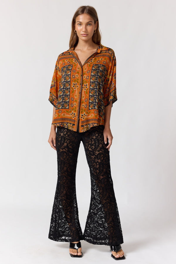 Woman wearing a patterned orange top and black lace pants on a white background