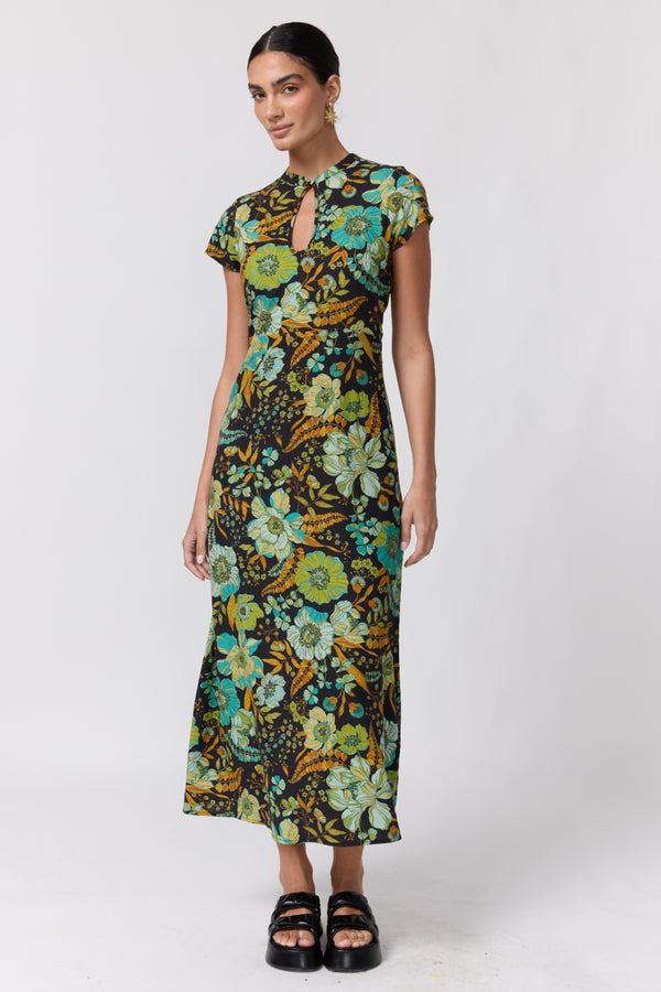 Nine Lives Bazaar Nostalgia Willa Maxi Dress. A woman modeling a short-sleeve midi dress with a vibrant floral print in green, yellow, and blue on a dark background, paired with black platform sandals, against a light gray studio backdrop.