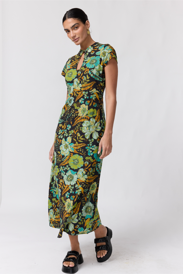 Nine Lives Bazaar Nostalgia Willa Maxi Dress. A woman modeling a short-sleeve midi dress with a vibrant floral print in green, yellow, and blue on a dark background, paired with black platform sandals, against a light gray studio backdrop.