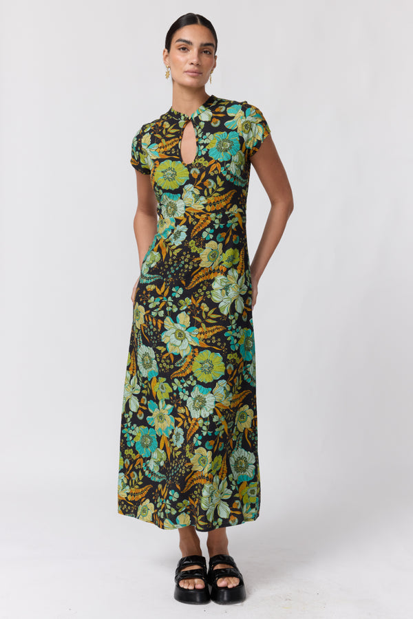 Nine Lives Bazaar Nostalgia Willa Maxi Dress. A woman modeling a short-sleeve midi dress with a vibrant floral print in green, yellow, and blue on a dark background, paired with black platform sandals, against a light gray studio backdrop.