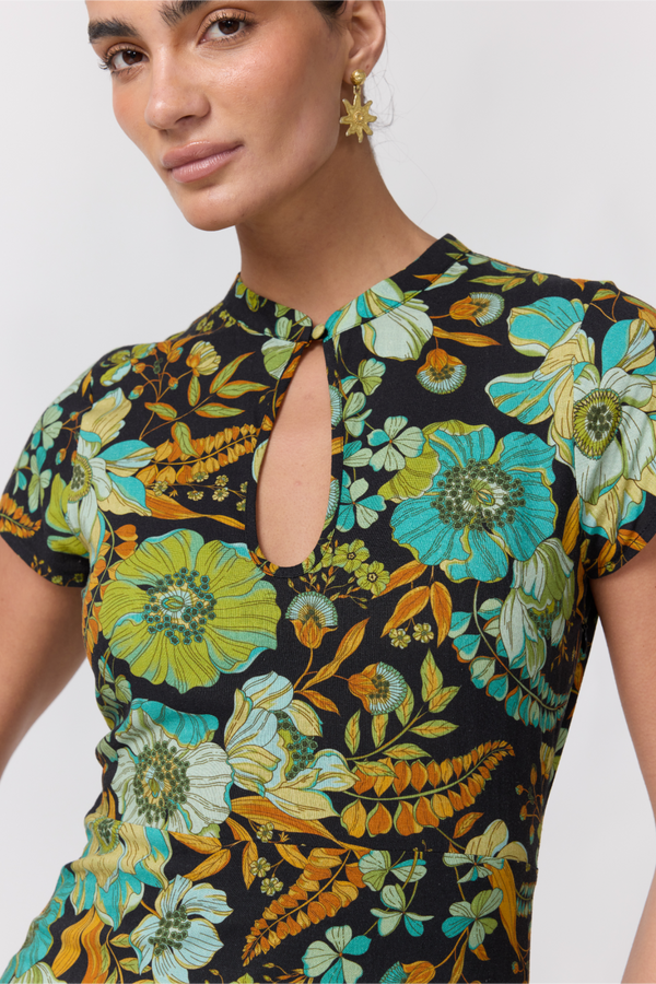 Nine Lives Bazaar Nostalgia Willa Maxi Dress. A close up of woman modeling a short-sleeve midi dress with a vibrant floral print in green, yellow, and blue on a dark background, paired with black platform sandals, against a light gray studio backdrop.