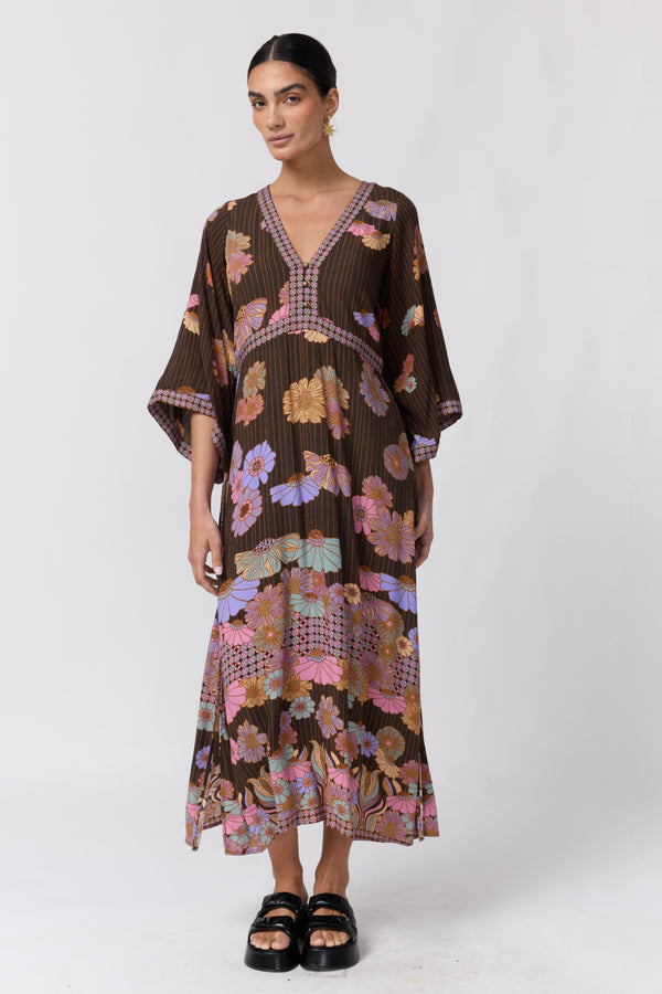 A woman modeling a brown pleated kaftan dress with a colorful floral and geometric print, paired with black platform sandals, against a plain light gray studio background.