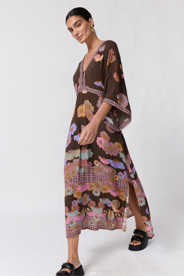A woman modeling a brown pleated maxi dress with a colorful floral and butterfly print, featuring wide three-quarter sleeves and a side slit, paired with black slide sandals against a light gray studio background.