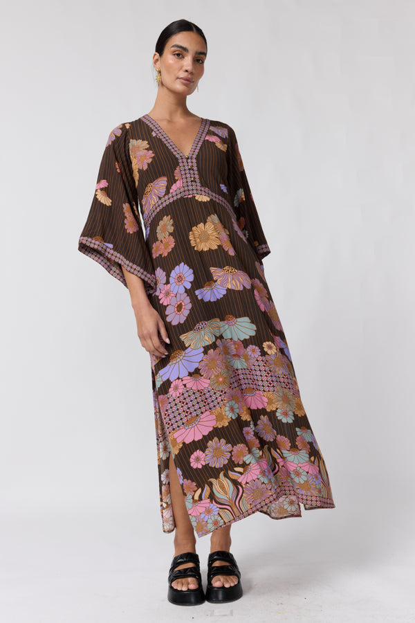 A woman modeling a brown kaftan dress with a vibrant multicolor floral and geometric print, paired with black platform sandals, against a plain white studio background.