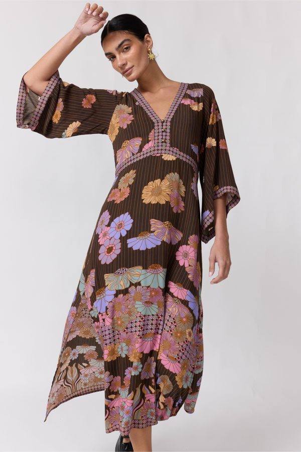 A woman models a brown kaftan dress with a colorful floral and butterfly print, featuring wide three-quarter sleeves and a V-neckline, set against a plain light gray background.