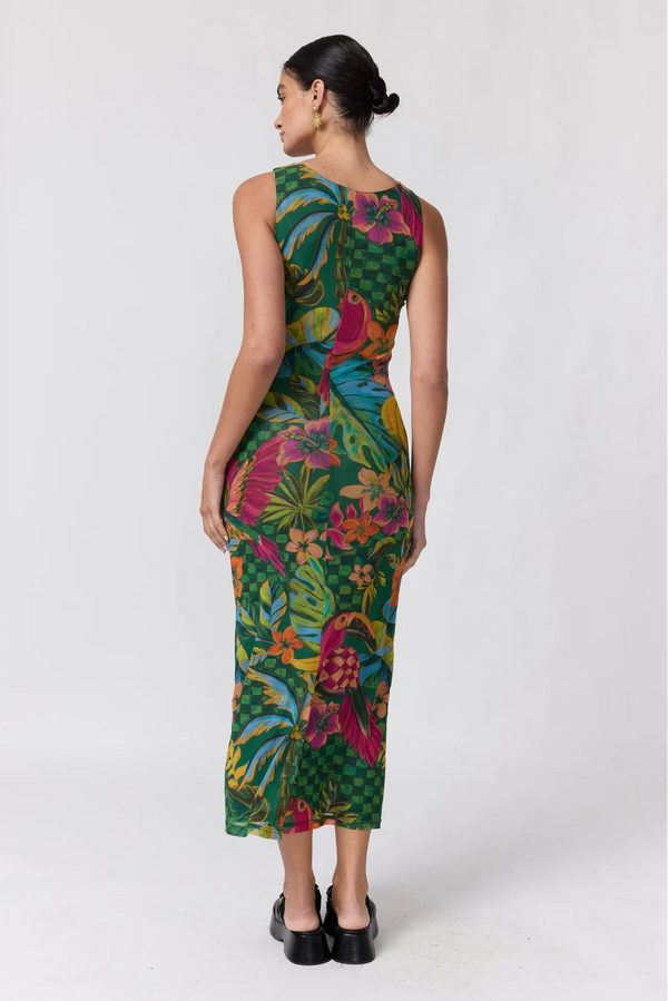 A woman stands with her back to the camera wearing a sleeveless, knee-length dress with a vibrant tropical floral and leaf print in green, pink, yellow, and blue, paired with black platform heels, against a plain white studio background.