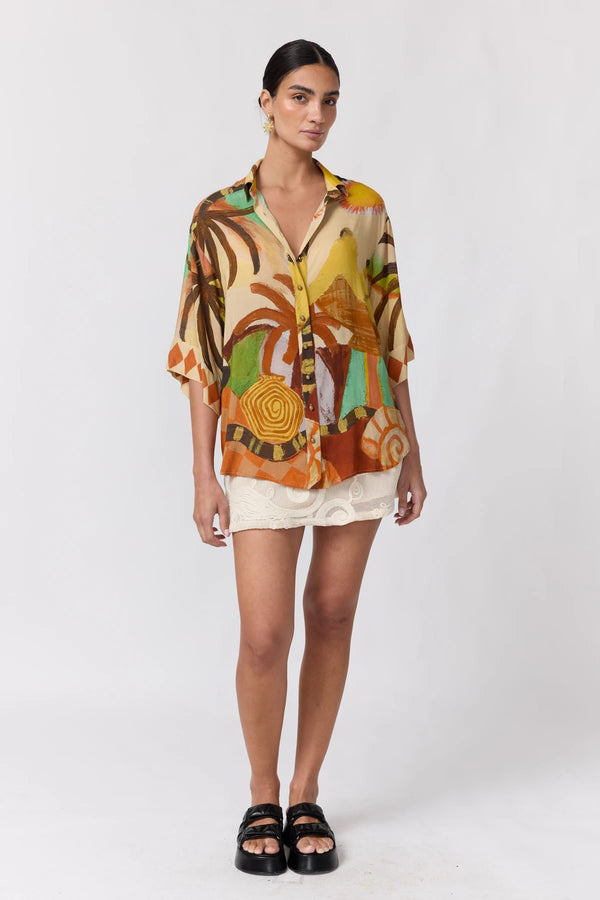 A model stands against a light gray studio background wearing a short-sleeve button-up shirt with a colorful abstract tropical-style print in orange, yellow, brown, green, and beige, paired with white shorts and black slide sandals.
