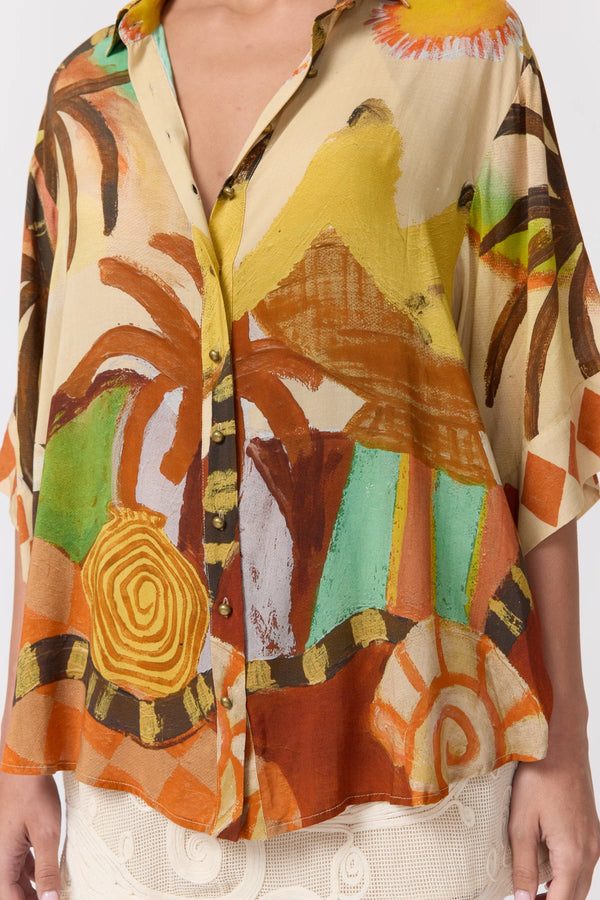A close-up of a woman wearing a loose, short-sleeve button-front blouse with a bold abstract tropical print in orange, yellow, brown, green, and cream.
