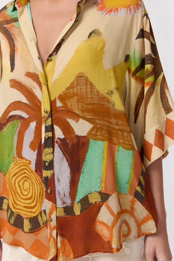 A close-up of a person wearing a loose short-sleeve button-up shirt with a bold abstract tropical print in beige, orange, brown, yellow, green, and white.