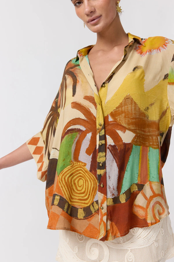 A woman wearing a short-sleeve button-up shirt with a vibrant abstract tropical print in orange, yellow, brown, green, and beige, paired with a white embroidered skirt against a plain white background.