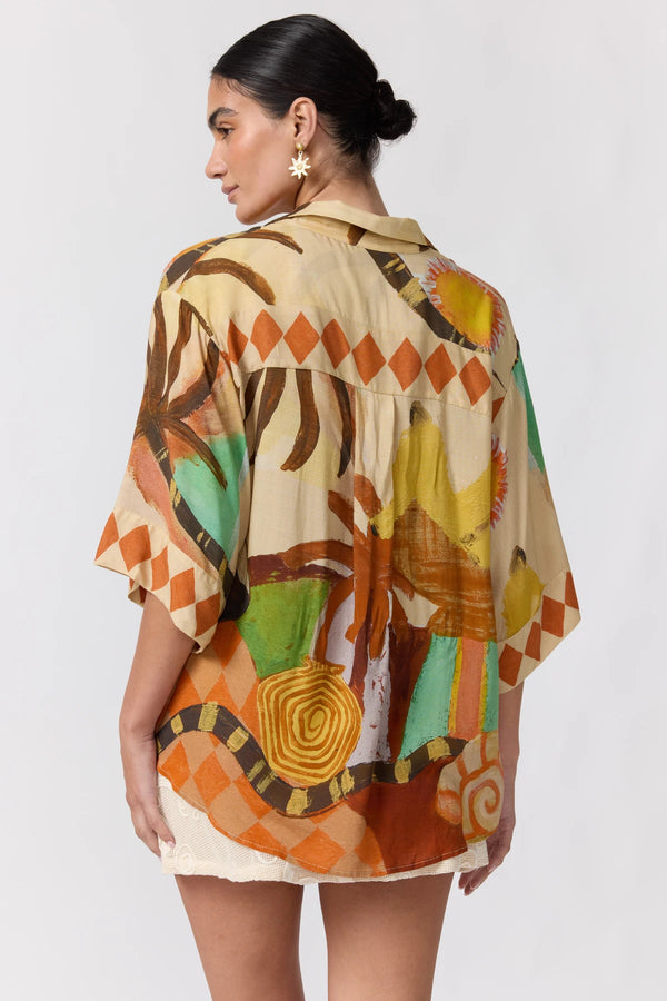 A woman shown from the back wearing a loose short-sleeve button-up shirt with a colorful abstract print in orange, green, yellow, brown, and beige, paired with white shorts against a light gray studio background.