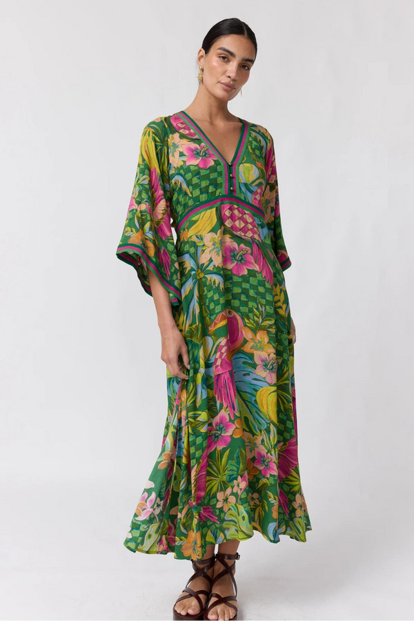 A woman modeling a vibrant green tropical print maxi dress with a V-neckline, three-quarter sleeves, and a colorful parrot motif, set against a plain light gray studio background.