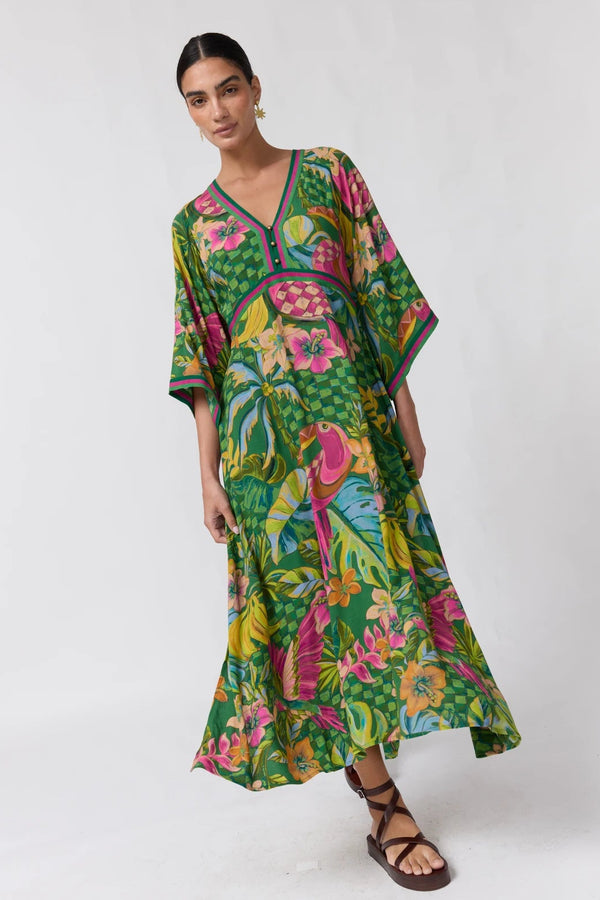 A woman modeling a vibrant green tropical print kaftan dress with pink flamingos, blue and yellow accents, and brown lace-up sandals against a plain white studio background.