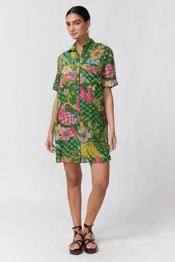 A woman modeling a short-sleeve button-up shirt dress with a vibrant tropical floral and bird print in green, pink, yellow, and blue, paired with brown strappy sandals against a plain white studio background.