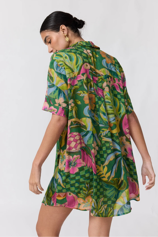 A woman modeling a short-sleeve, collared mini dress with a vibrant tropical print featuring green palm leaves, pink flamingos, and yellow accents, photographed against a plain light gray background.