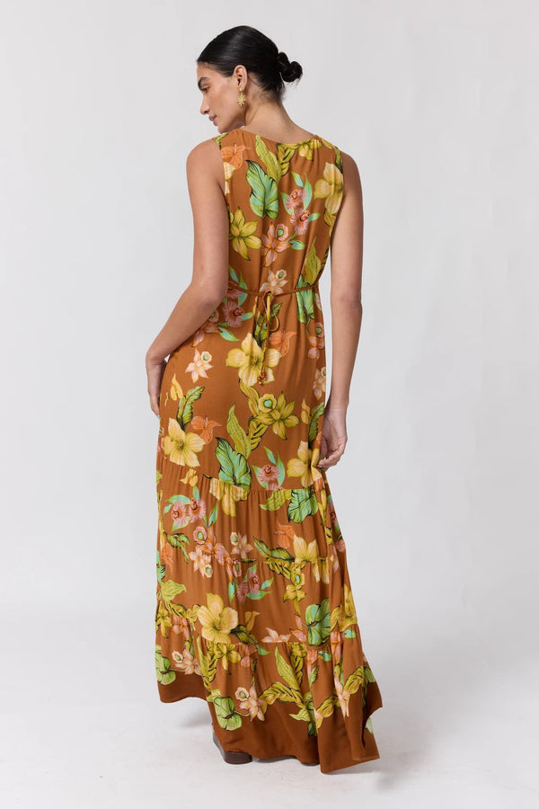 A woman modeling a sleeveless, tiered maxi dress from the back, featuring a brown base with a vibrant yellow, green, and orange floral pattern, set against a plain light gray studio background.