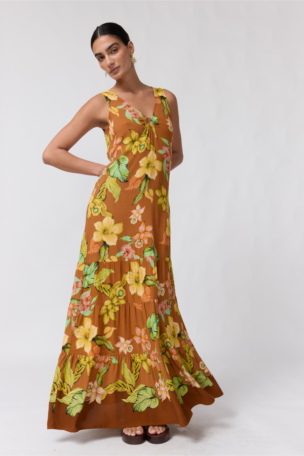 A woman modeling a sleeveless, tiered maxi dress with a brown base and a vibrant yellow, green, and orange floral print, set against a plain light gray studio background.