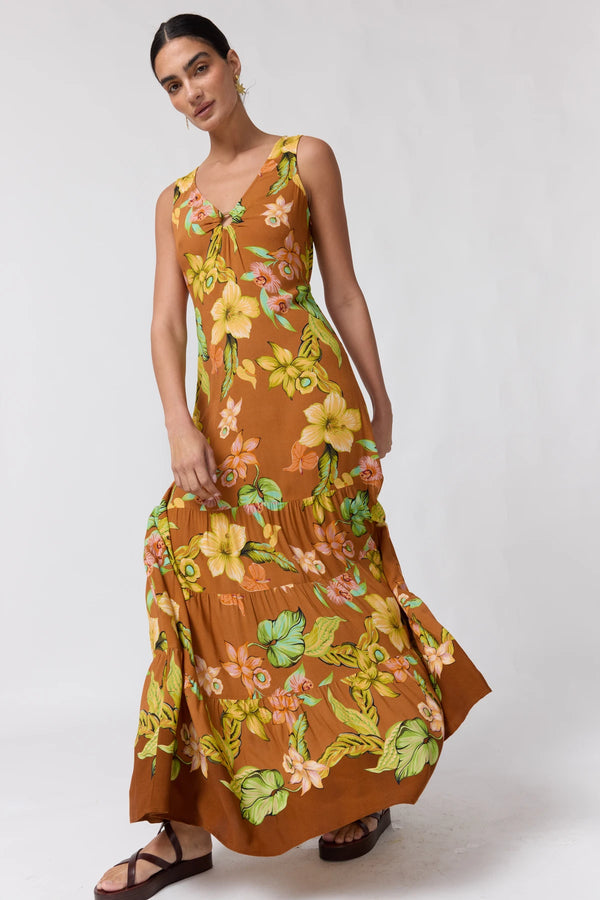 A woman modeling a sleeveless, V-neck maxi dress with a brown base and a vibrant yellow, green, and orange floral print, paired with dark brown strappy sandals against a light gray studio background.