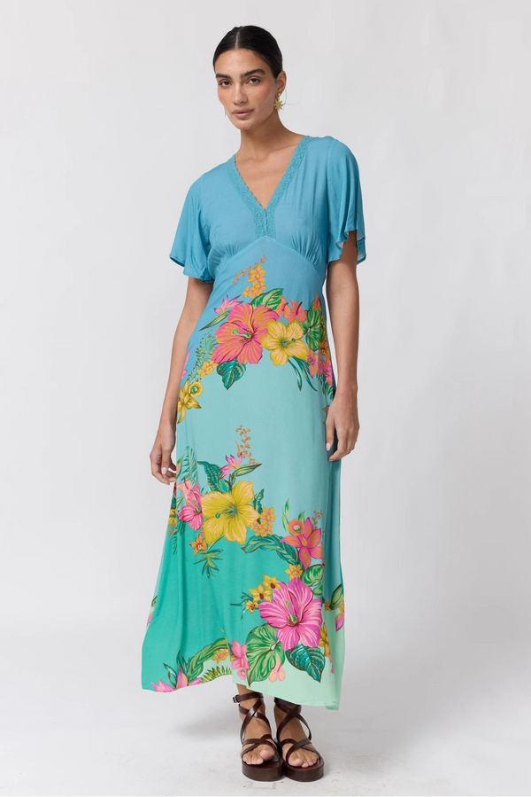 A woman modeling a short-sleeve maxi dress with a vibrant tropical floral print. The dress features a turquoise blue bodice and upper skirt that fades into a lighter aqua green lower skirt, decorated with large pink, yellow, and orange hibiscus flowers and green leaves.