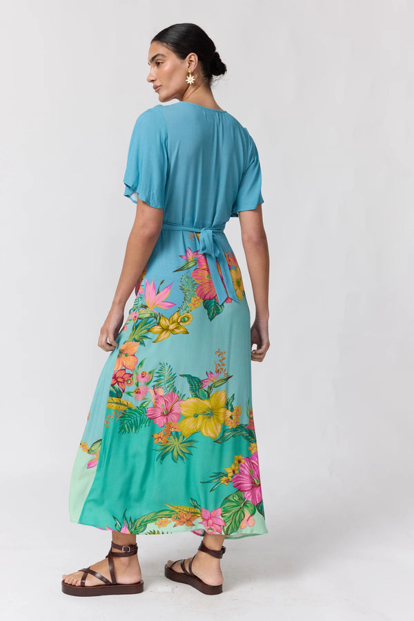 A woman modeling a turquoise maxi dress with a vibrant tropical floral print on the lower half, shown from a rear three-quarter angle against a light gray studio background.