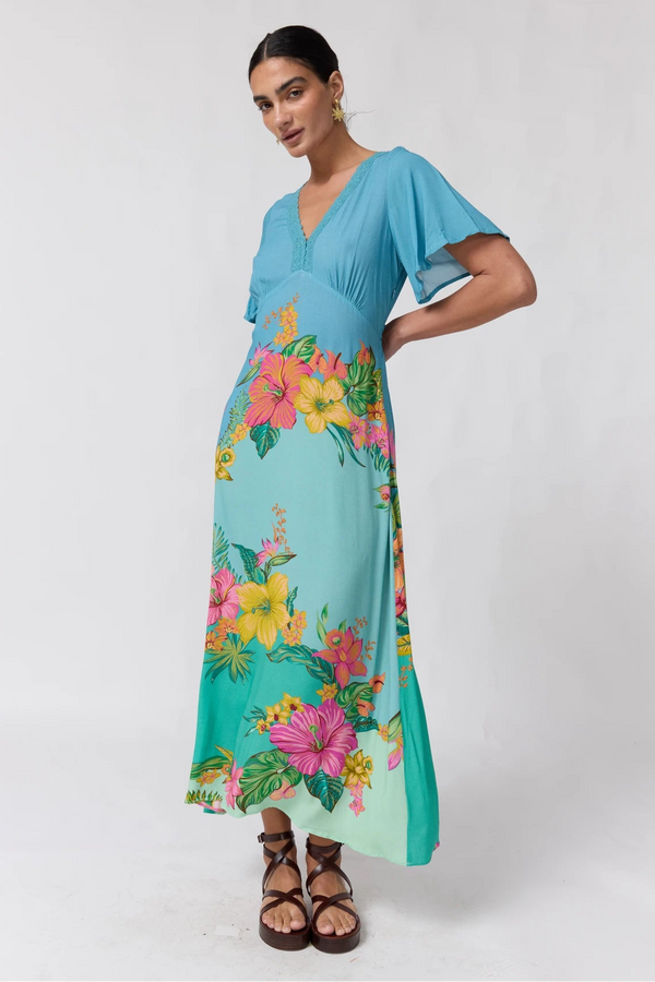 A woman modeling a light blue maternity maxi dress with a vibrant tropical floral print in pink, yellow, and green, paired with brown strappy sandals against a plain white studio background.
