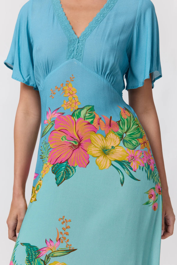 A woman wearing a light blue V-neck dress with short flutter sleeves and a colorful tropical floral print featuring pink hibiscus, yellow flowers, and green leaves.