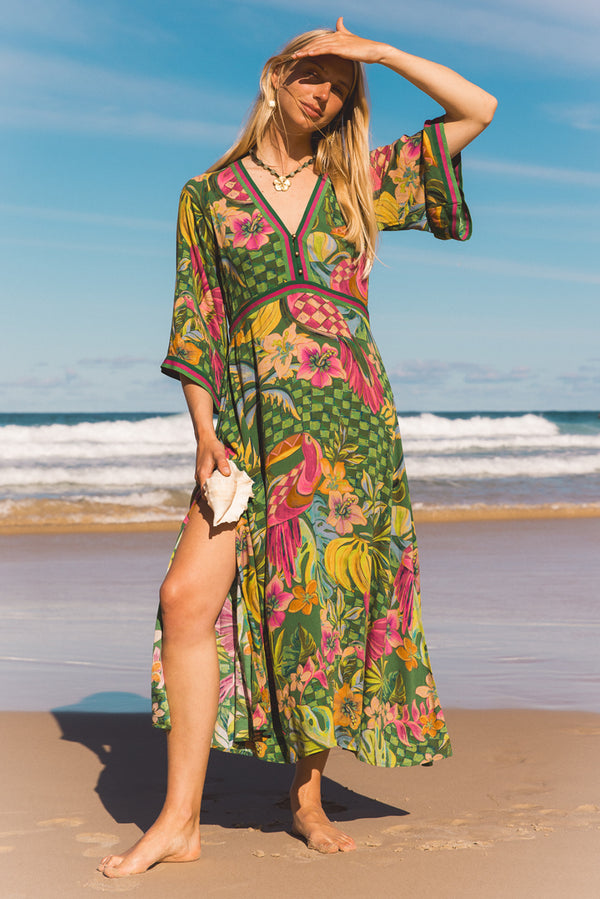 A woman stands on a sandy beach wearing a vibrant tropical print maxi dress with pink flamingos, toucans, and green foliage on a blue and green background. She is barefoot, holding a small white clutch, with the ocean and blue sky behind her. Location is Dreamtime Beach, Fingal Head, NSW.