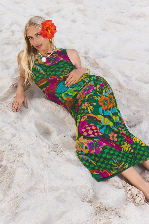 A woman reclining on white sand wearing a vibrant green tropical floral maxi dress with a red hibiscus flower in her hair.