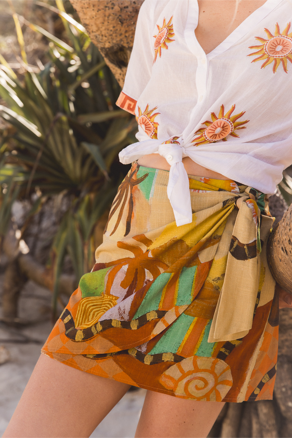 A close-up lifestyle shot of a model wearing a white sheer tie-front blouse with orange sun motifs and a multicolor abstract print wrap skirt in orange, beige, and green. Location is Dreamtime Beach, Fingal Head, NSW.