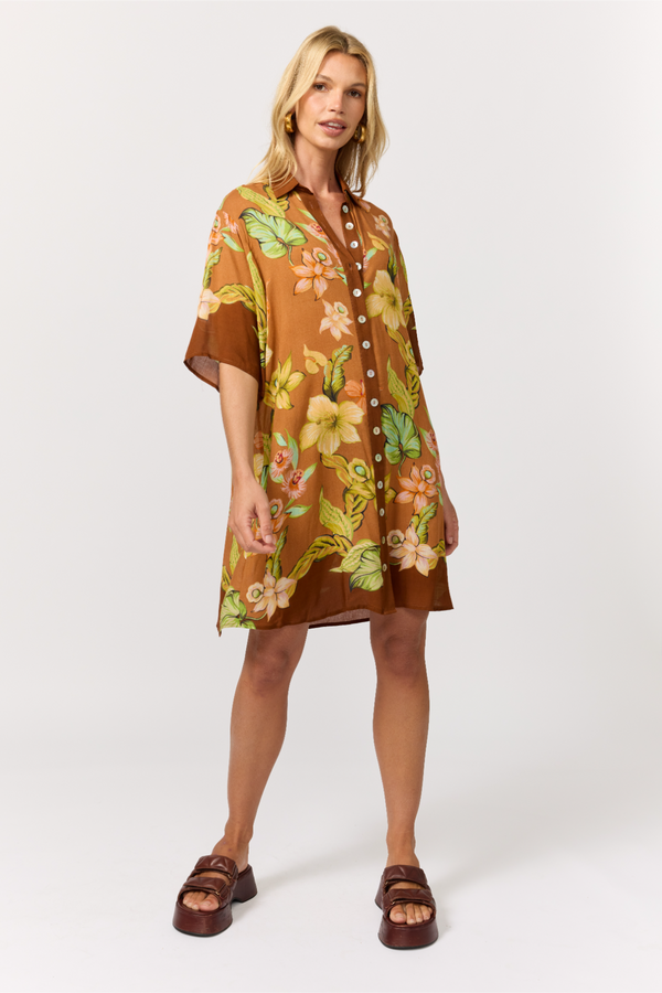 A woman modeling a brown short-sleeve button-front shirt dress with a tropical floral print in yellow, green, and peach, paired with brown slide sandals, against a light gray studio background.