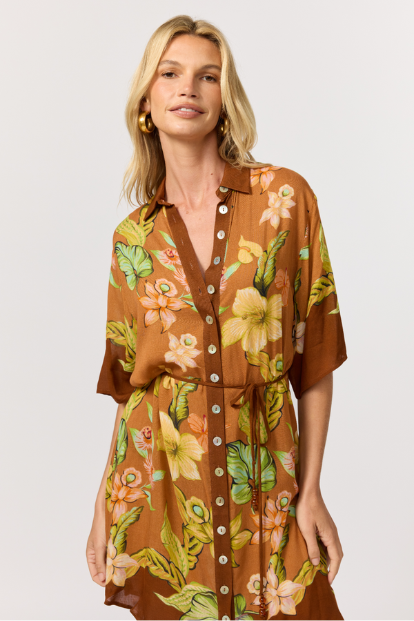 A woman modeling a brown short-sleeve button-front shirt dress with a vibrant tropical floral print in yellow, orange, and green, set against a plain light gray studio background.