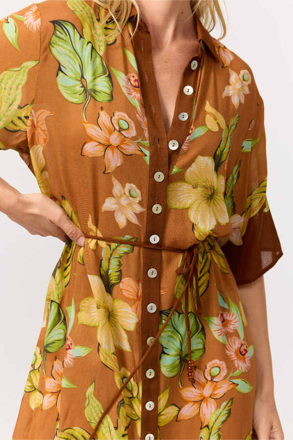 A close-up of a woman wearing a brown button-front shirt dress with a tropical floral print in green, yellow, and orange. The dress has a tie waist and is shown against a plain light background.
