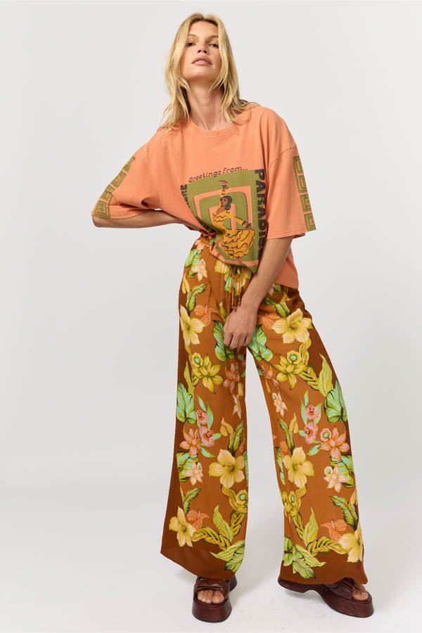 A model wearing a matching two-piece outfit consisting of a peach graphic t-shirt and brown wide-leg pants with a yellow and green floral print, photographed against a light gray studio background.