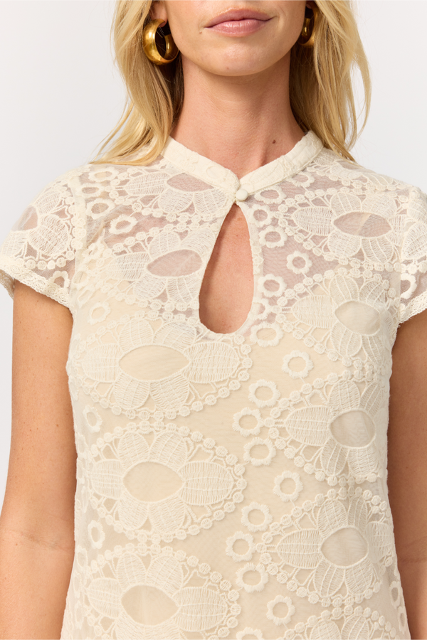 A close-up of a woman wearing a cream-colored lace top with a keyhole cutout at the collar and floral lace patterns. She is wearing large gold hoop earrings against a plain light background.
