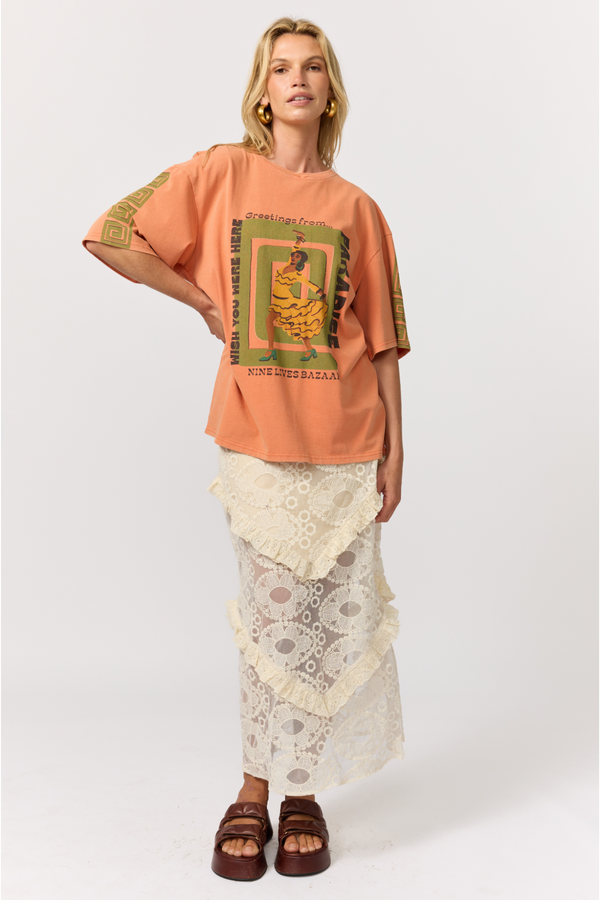 A model stands against a light gray studio background wearing an oversized peach-orange graphic T-shirt with green and yellow print, a cream lace tiered skirt, and brown strappy sandals.