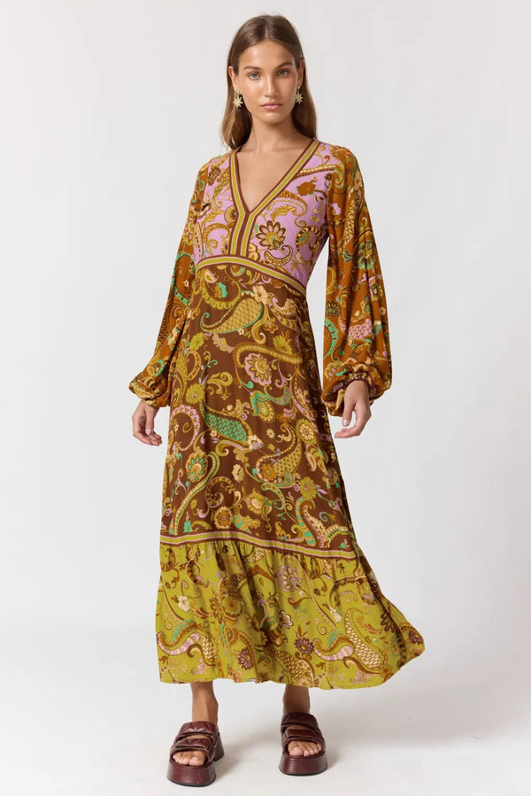A woman modeling a long-sleeved maxi dress with a complex paisley and floral pattern in mustard yellow, brown, purple, and green, paired with brown platform sandals against a plain white studio background.