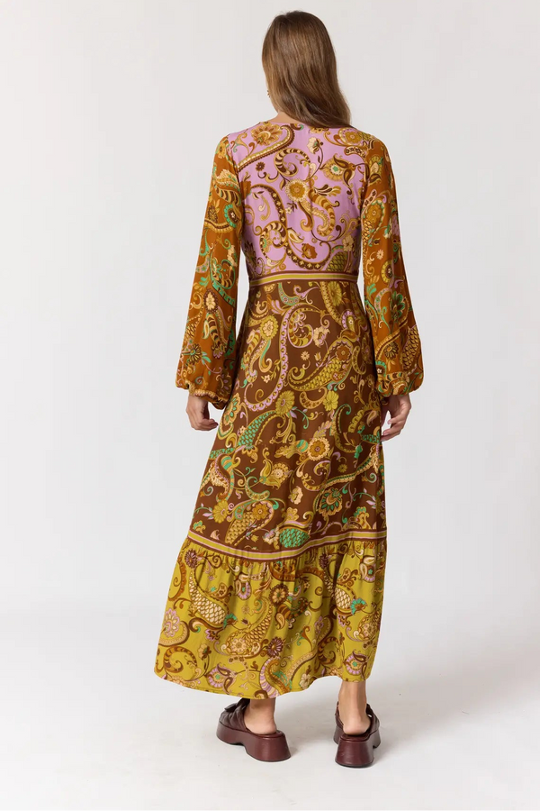 A woman viewed from the back wearing a long-sleeved maxi dress with a complex paisley and floral pattern in brown, yellow, and lavender, paired with dark brown shoes, against a plain white studio background.
