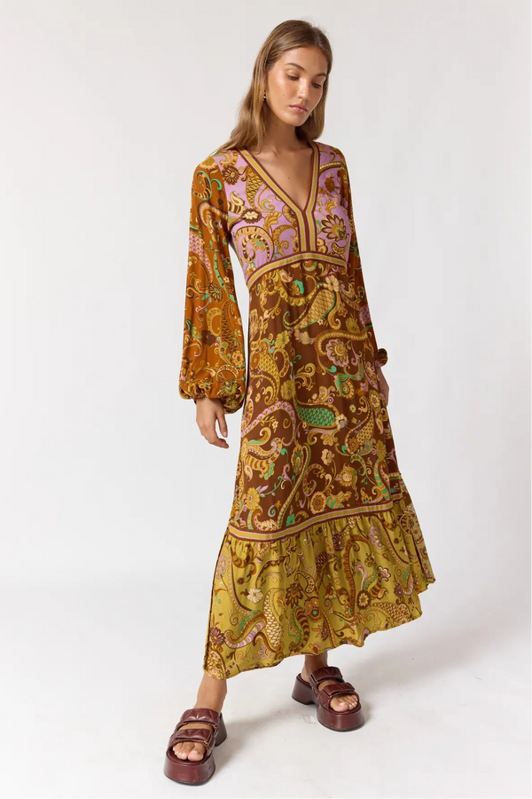A woman modeling a long-sleeved bohemian maxi dress with an intricate paisley and floral pattern in brown, gold, purple, and green, paired with brown platform sandals against a plain white studio background.