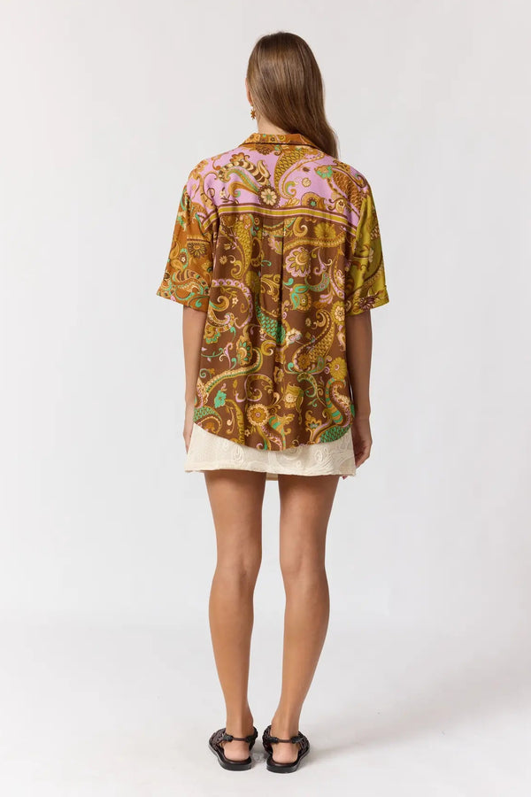 A woman viewed from the back wearing a short-sleeve button-up shirt with a colorful paisley print in brown, gold, pink, and green, paired with a light beige mini skirt and black sandals, against a plain white studio background.