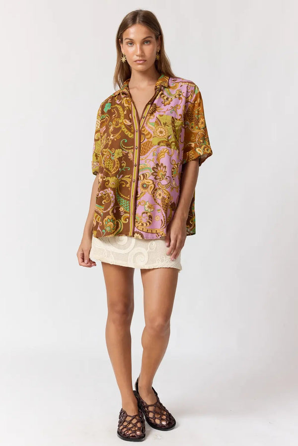 A model stands against a light gray studio background wearing a short-sleeve button-up shirt with an ornate multicolor paisley print in gold, brown, pink, and green, paired with a short cream skirt and dark strappy flat sandals.