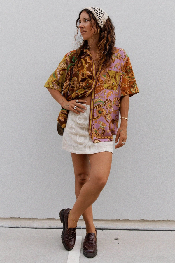 A woman stands against a plain light gray wall wearing a short-sleeve button-up shirt with a colorful paisley print in orange, yellow, pink, and brown, paired with a white mini skirt, brown loafers, and a white patterned headband.