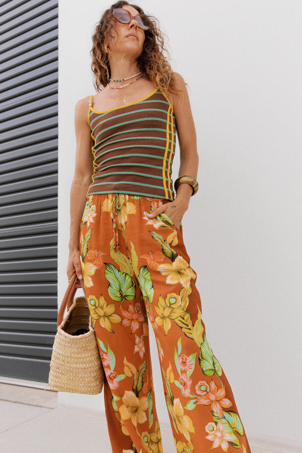 A woman models a two-piece outfit featuring a brown and green striped ribbed tank top with yellow trim, paired with wide-leg orange trousers with a yellow, green, and pink floral print. She is holding a woven straw bag and posing against a white wall with a dark gray slatted screen.