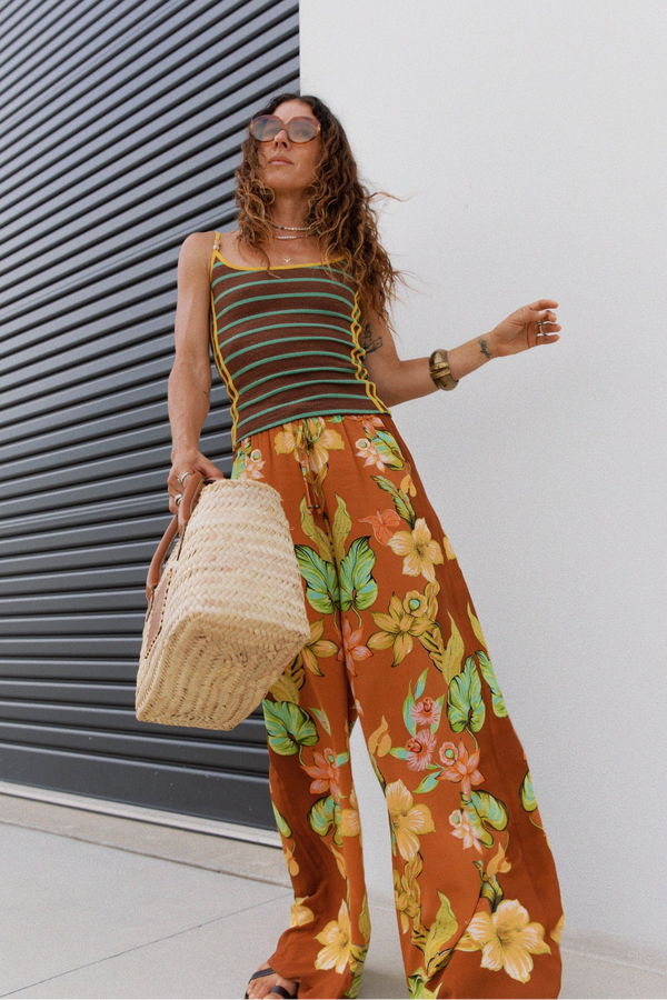 A woman wearing a brown and orange striped camisole top and matching wide-leg floral pants in rust orange, holding a woven straw tote bag, posing in front of a white wall and a dark gray metal shutter.