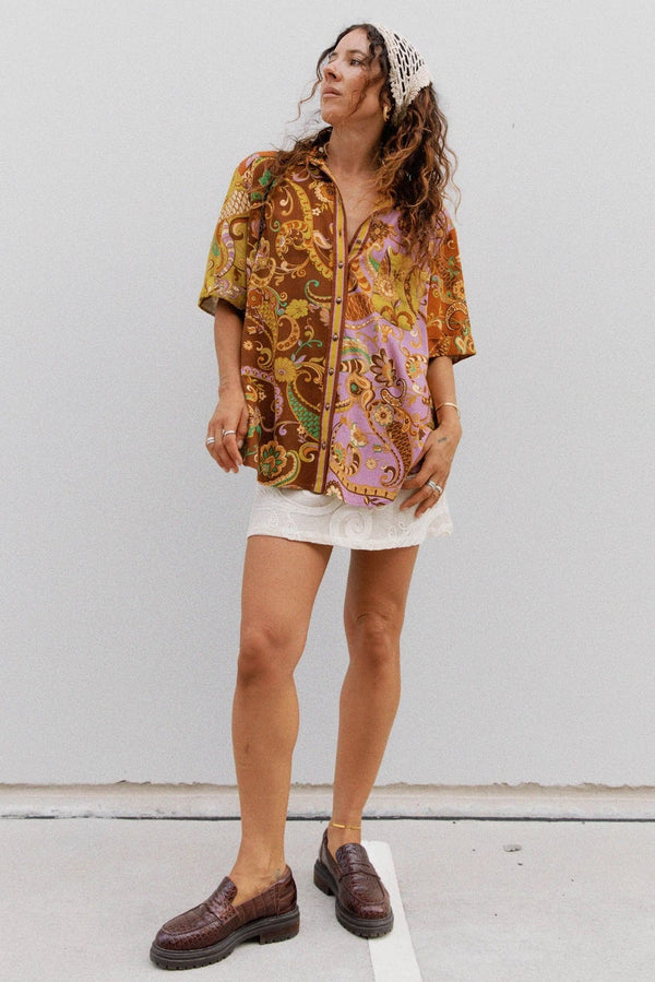 A woman stands against a plain light gray wall wearing a loose short-sleeve button-up shirt with an ornate multicolor paisley and floral print in gold, brown, pink, and green tones, paired with a white skirt and dark brown loafers.