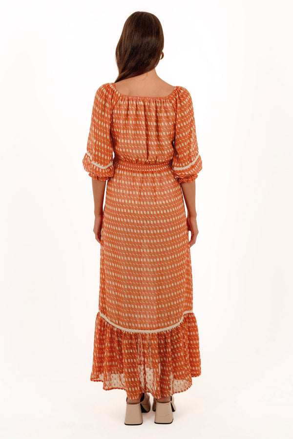 A woman viewed from the back wearing a rust-orange and cream patterned maxi dress with puff sleeves and a tiered ruffle hem, paired with beige block-heel sandals, against a plain white studio background.