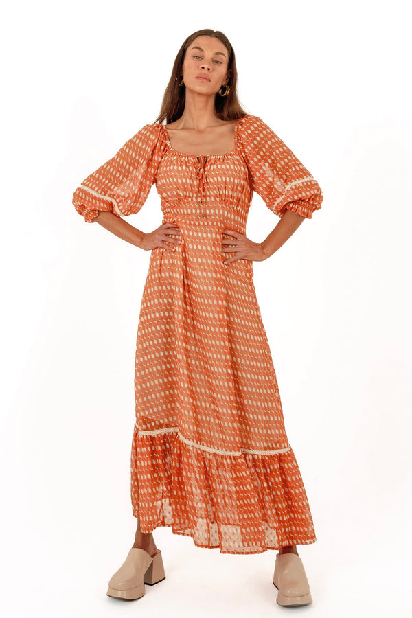A woman modeling a long-sleeved orange and white patterned maxi dress with puff sleeves, a square neckline, and a tiered ruffle hem, paired with beige block-heel mules against a plain white background.