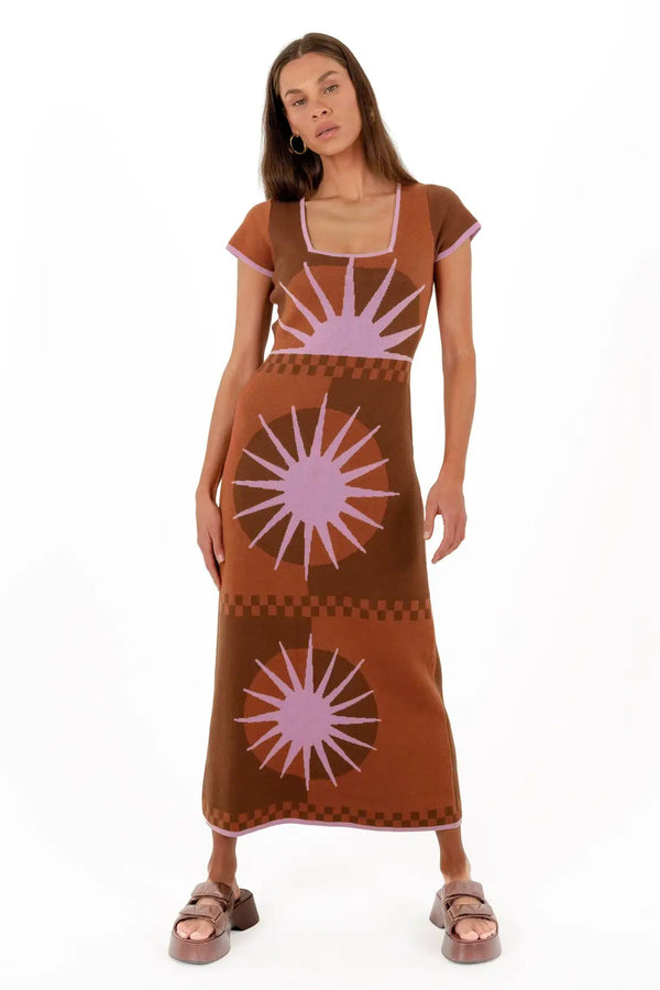 A woman modeling a brown maxi dress with a light purple geometric sunburst pattern, styled with brown slide sandals against a plain white background.