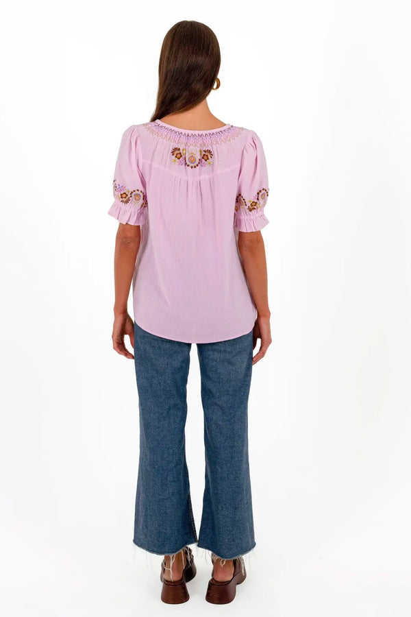 A woman viewed from the back wearing a light pink short-sleeve blouse with embroidered details on the back neckline and sleeves, paired with blue flared jeans and brown shoes, against a plain white background.